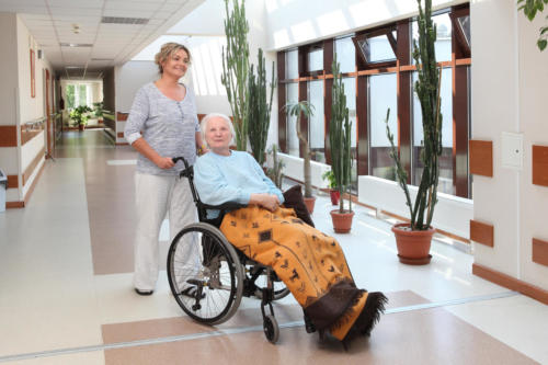 zdjęcie opiekunka prowadząca wózek inwalidzki z mieszkanką domu. widoczny jest przestronny korytarz i dużo otaczającej je zieleni z kwiatów doniczkowych.